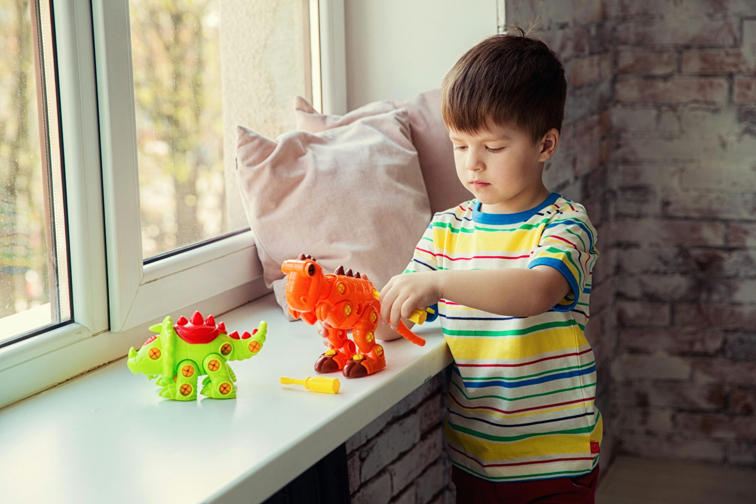 Young boy playing with colorful dinosaur toys on a windowsill, wearing a striped shirt, indoors.
